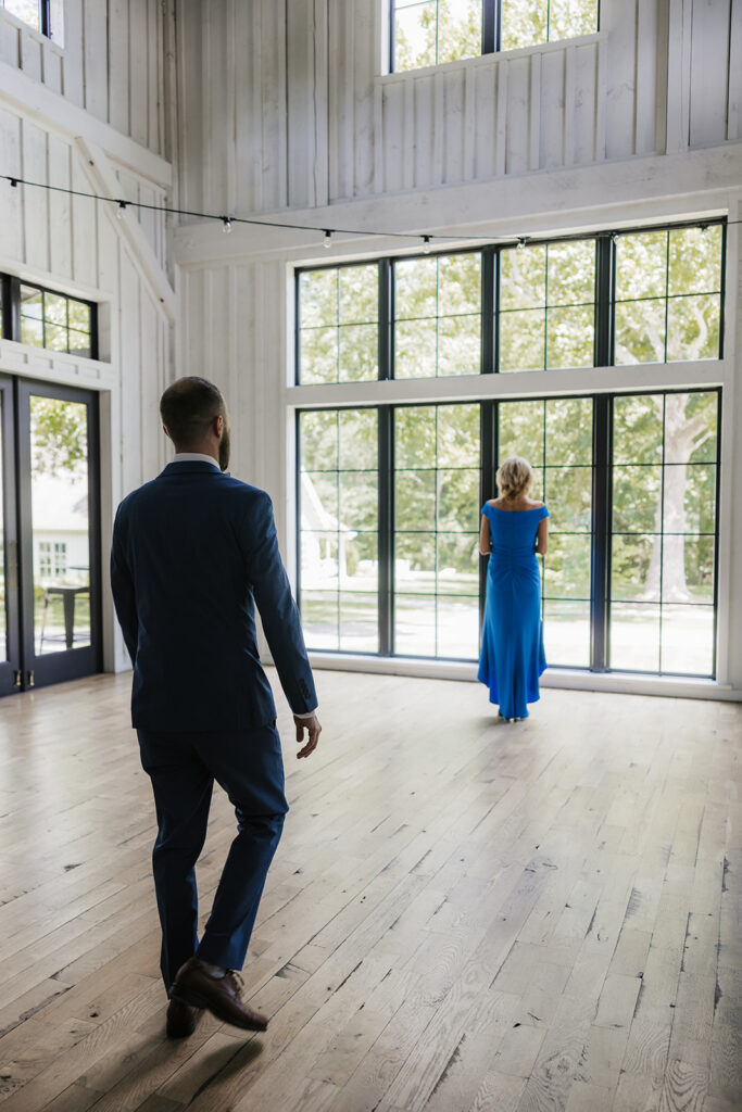 A man in a suit walks toward a woman in a blue dress standing by large windows in a bright, empty room with wooden floors.
