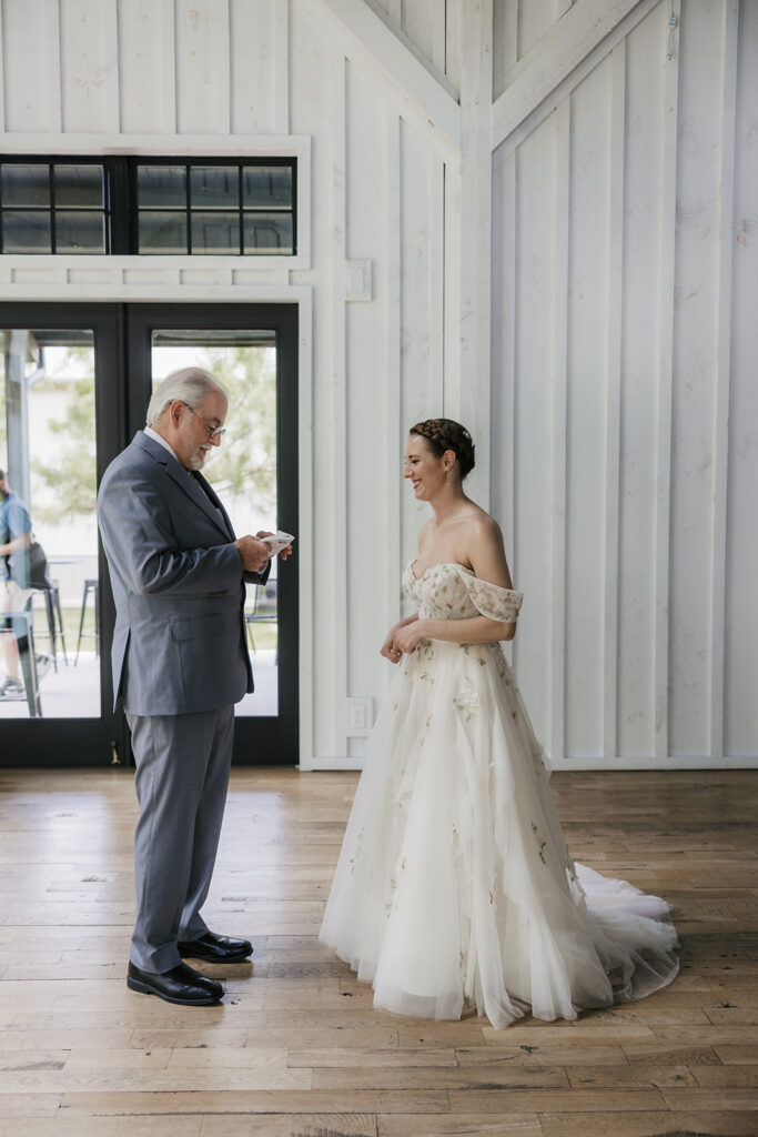 A man in a suit reads from a card while standing across from a woman in a wedding dress inside a bright, wooden-paneled room.