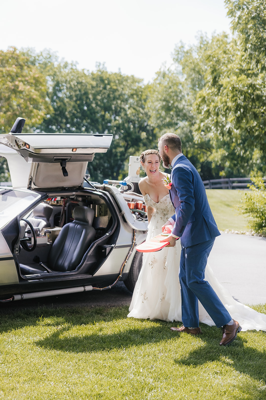 A bride and groom stand next to a DeLorean with open gull-wing doors on a grassy area, with trees in the background.
