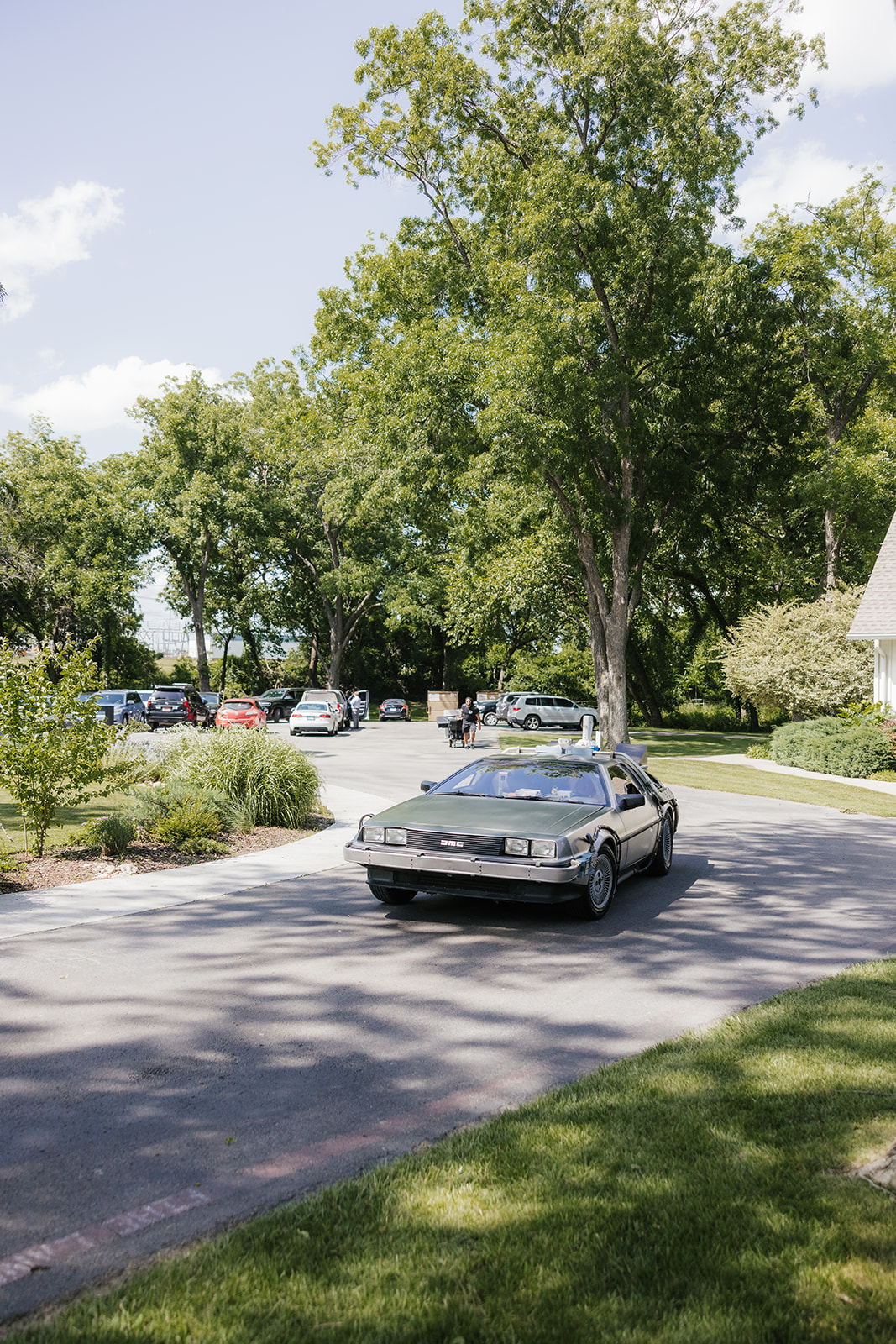 A silver DeLorean sports car with its gull-wing doors closed is parked on a sunlit road surrounded by trees and greenery.