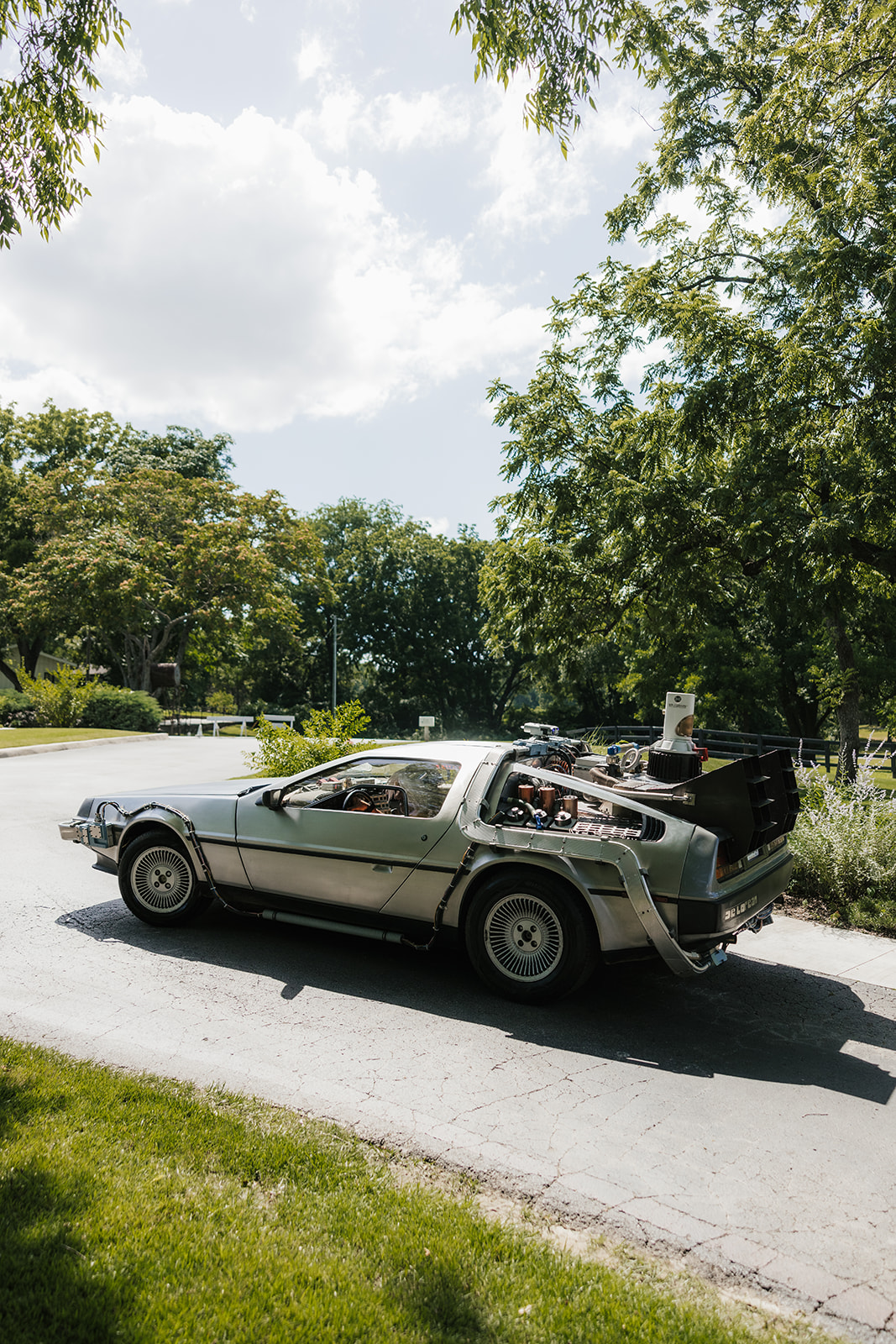 A silver DeLorean sports car with its gull-wing doors closed is parked on a sunlit road surrounded by trees and greenery.