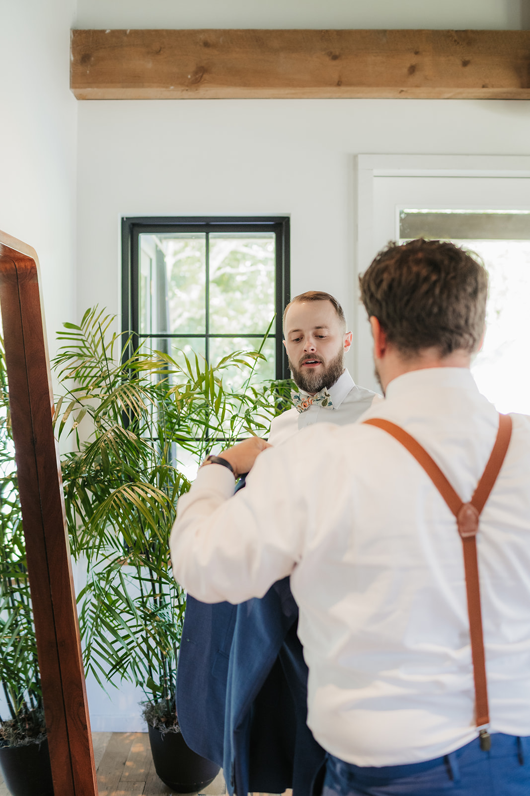 A man in a blue suit stands in front of a large mirror, adjusting his hands, with a plant to the right and shoes on the floor nearby.