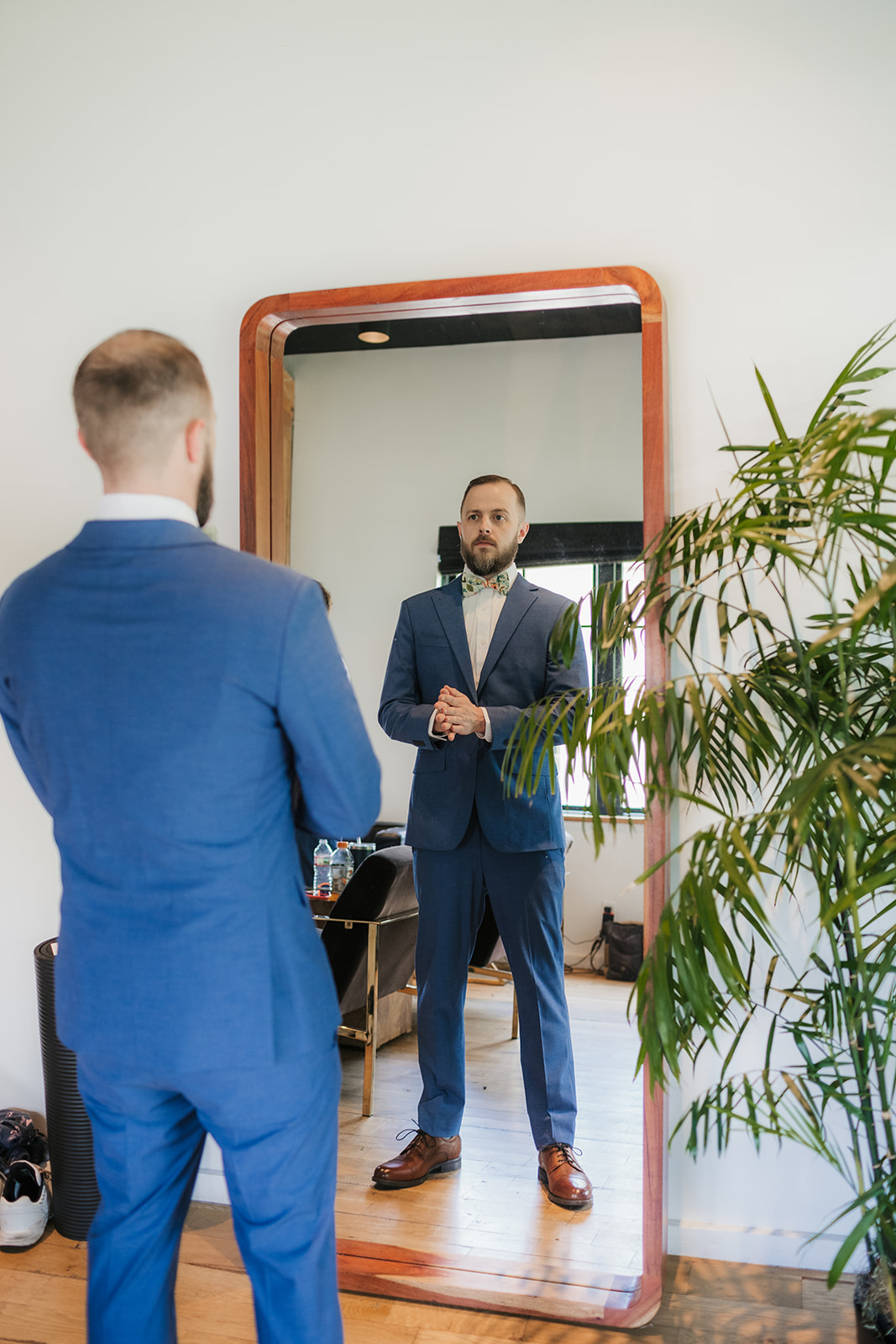 A man in a blue suit stands in front of a large mirror, adjusting his hands, with a plant to the right and shoes on the floor nearby.