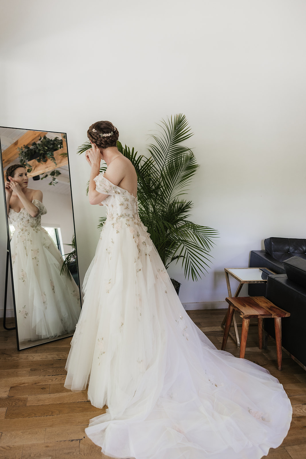 A bride in a white, off-the-shoulder floral gown puts on earrings, standing in front of a leafy plant.