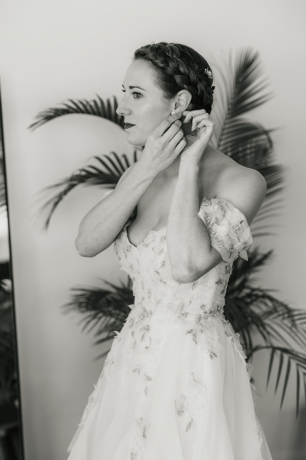 A bride in a white, off-the-shoulder floral gown puts on earrings, standing in front of a leafy plant.