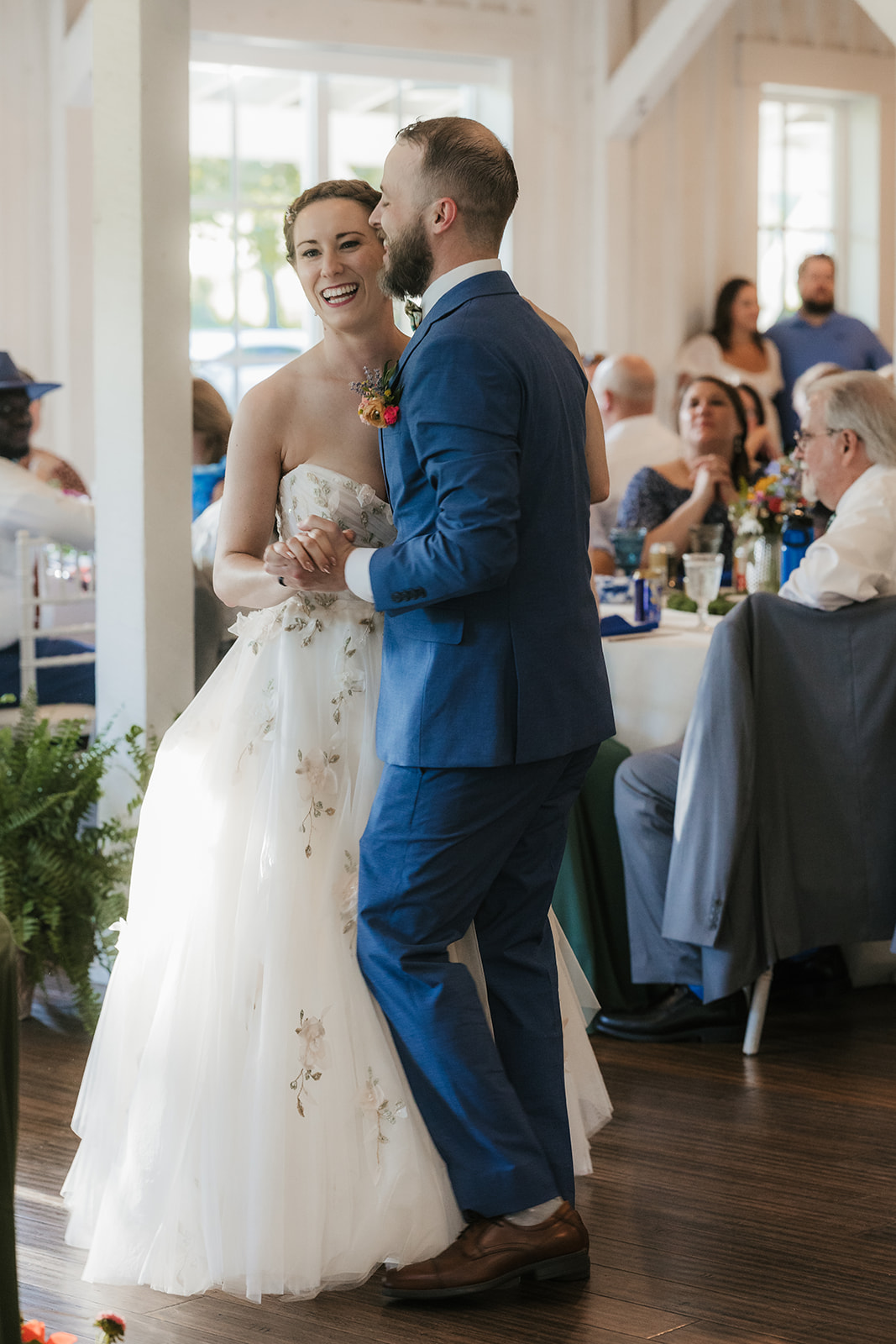 A bride in a white dress and a groom in a blue suit dance together at their wedding reception, surrounded by seated guests.