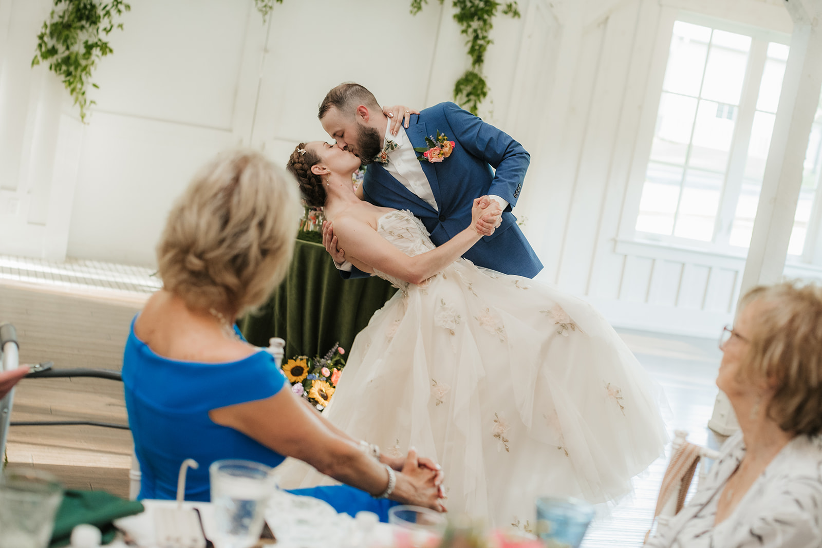 A bride in a white dress and a groom in a blue suit dance together at their wedding reception, surrounded by seated guests.