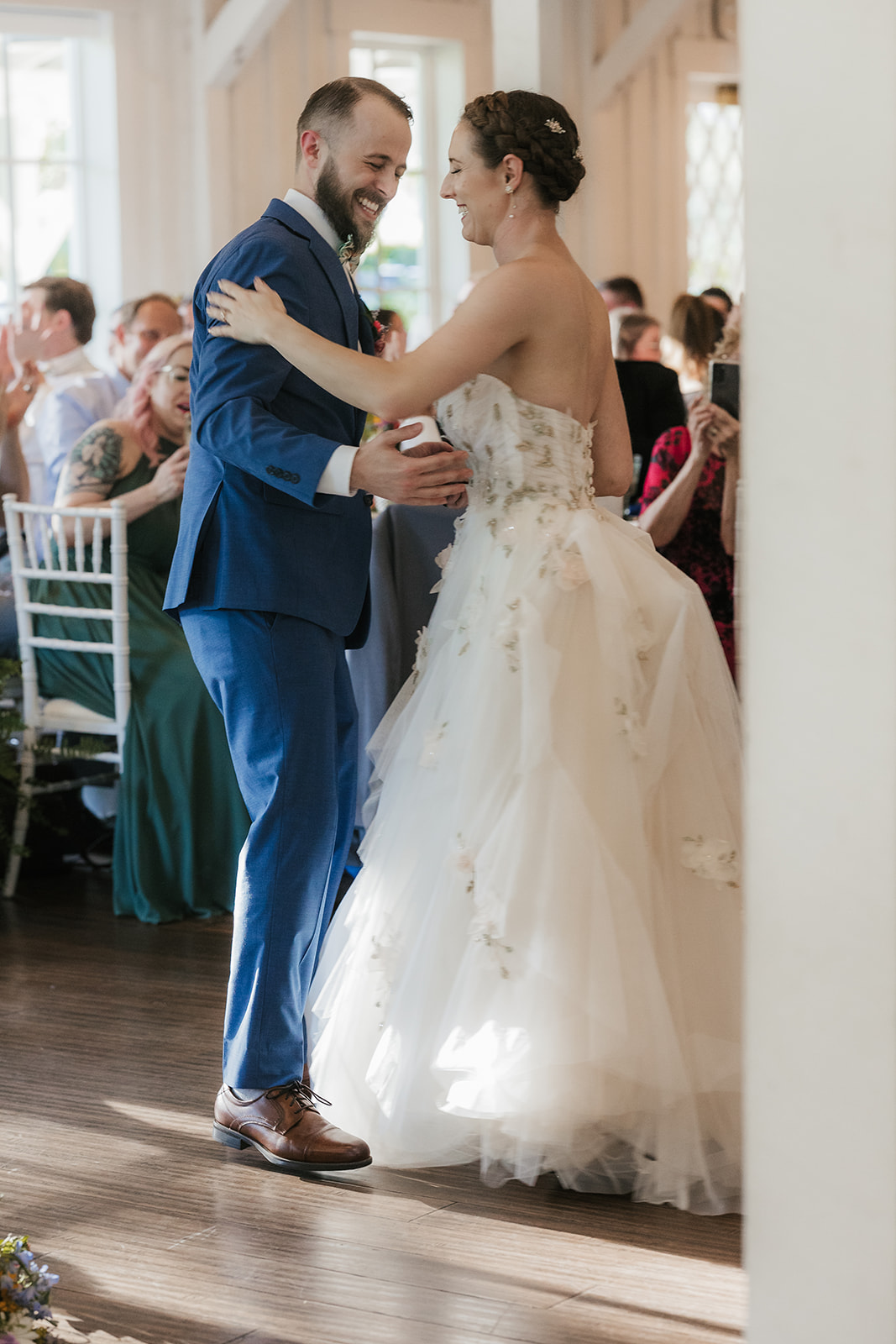 A bride in a white dress and a groom in a blue suit dance together at their wedding reception, surrounded by seated guests.