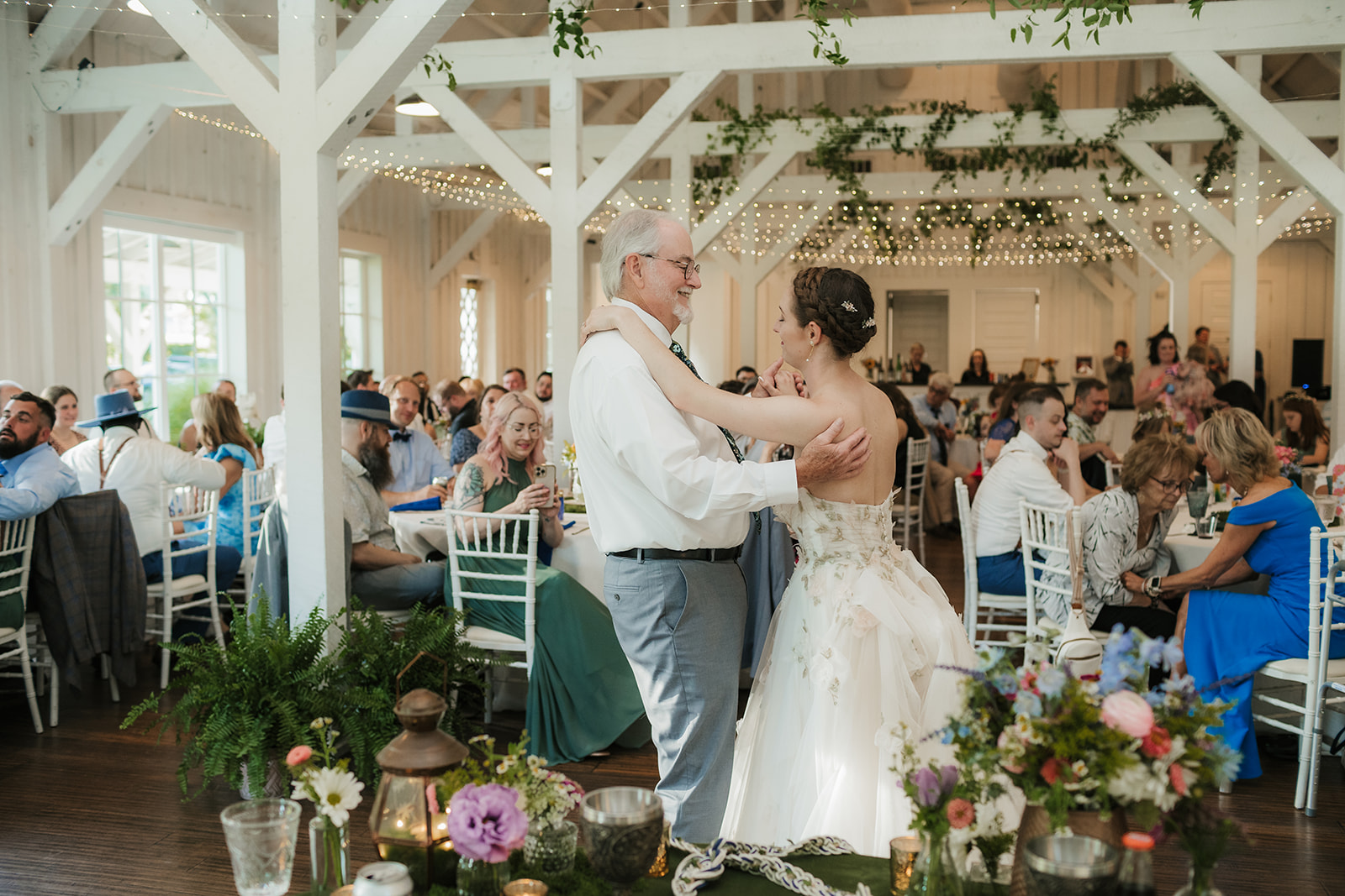 A bride in a white dress and a groom in a blue suit dance together at their wedding reception, surrounded by seated guests.