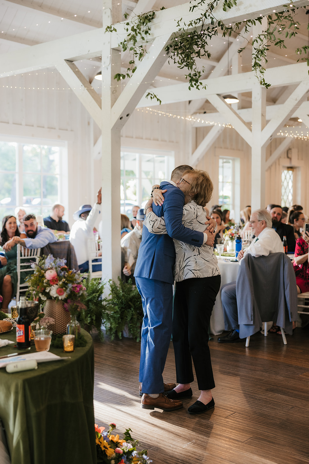 A bride in a white dress and a groom in a blue suit dance together at their wedding reception, surrounded by seated guests.