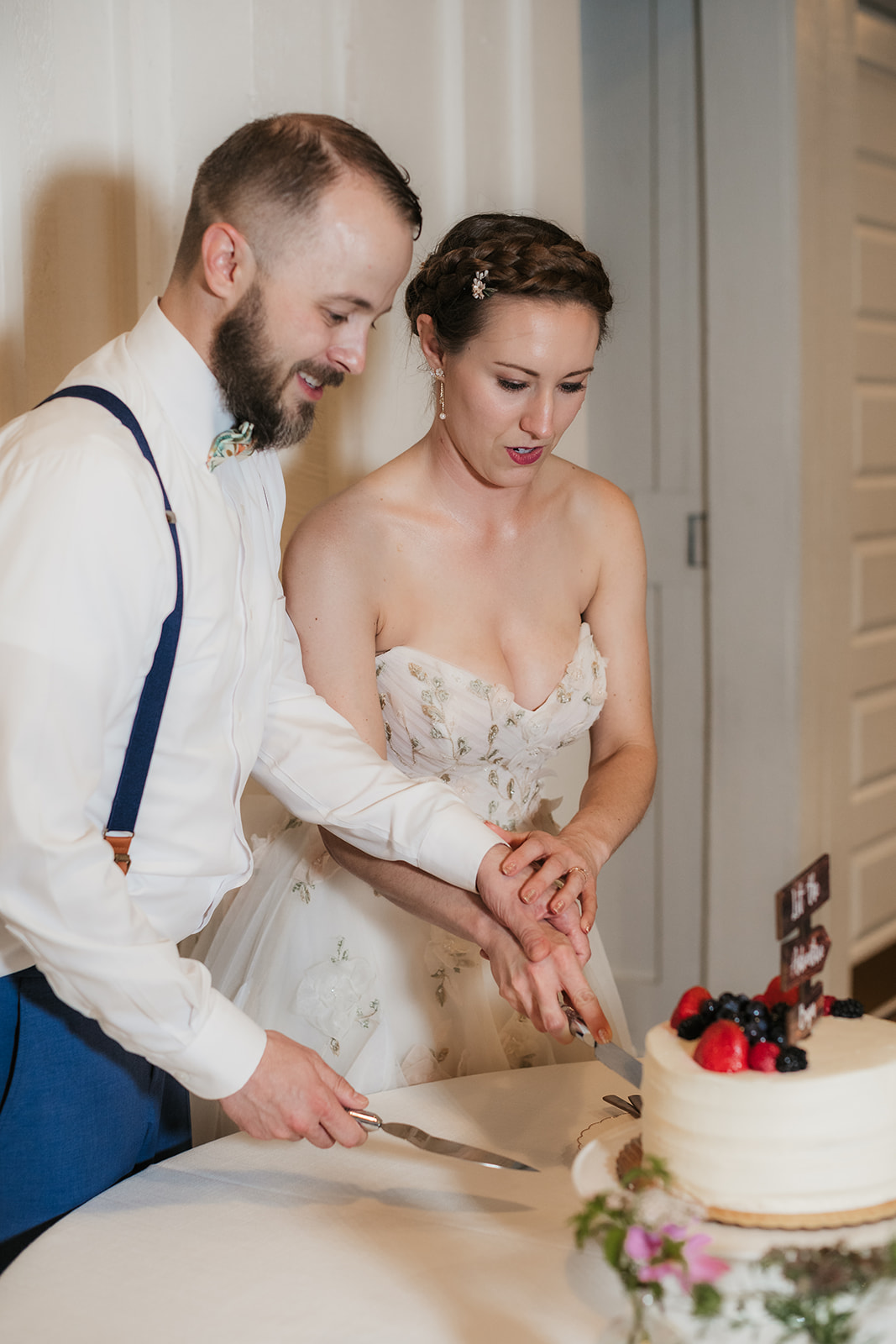 A bride and groom cut a white-tiered cake topped with berries at a table; the bride appears to be making a surprised expression.