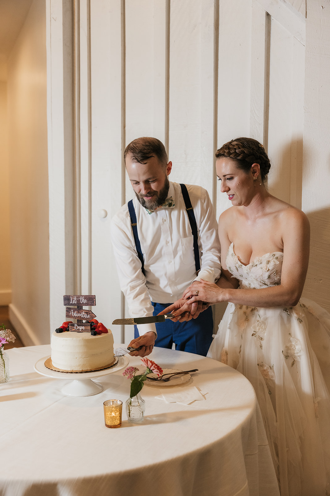 A bride and groom cut a white-tiered cake topped with berries at a table; the bride appears to be making a surprised expression.