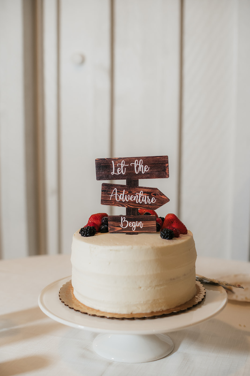 A white frosted cake topped with berries and a wooden sign that reads “Let the Adventure Begin” sits on a white cake stand.