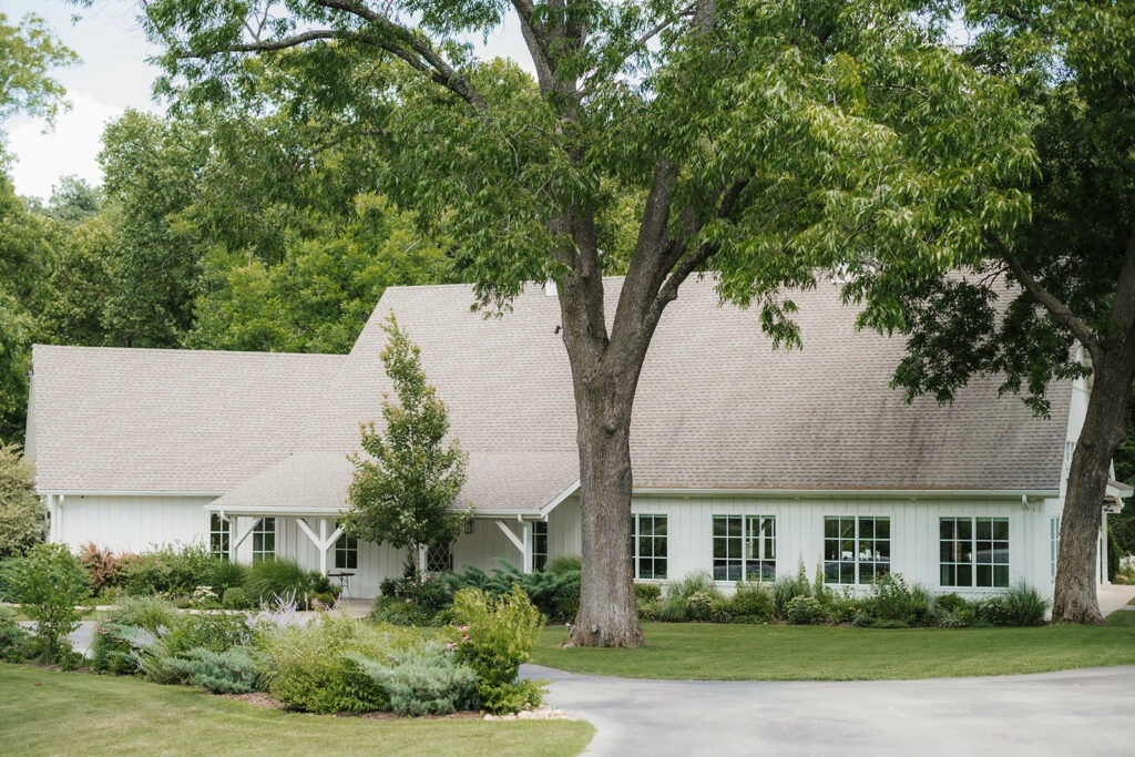 Single-story white house with a large, sloped roof, surrounded by trees and greenery, with a curved driveway in front.
