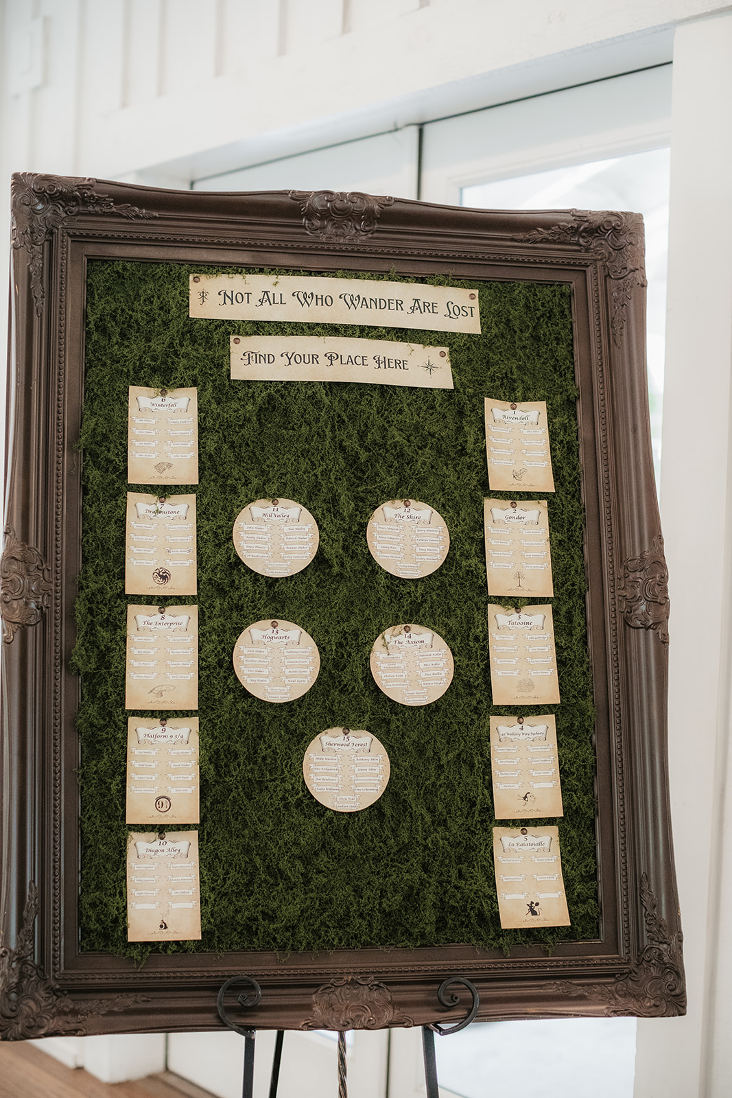 A framed seating chart display with individual cards arranged in rows and circles, mounted on a green moss background, stands indoors by a window.