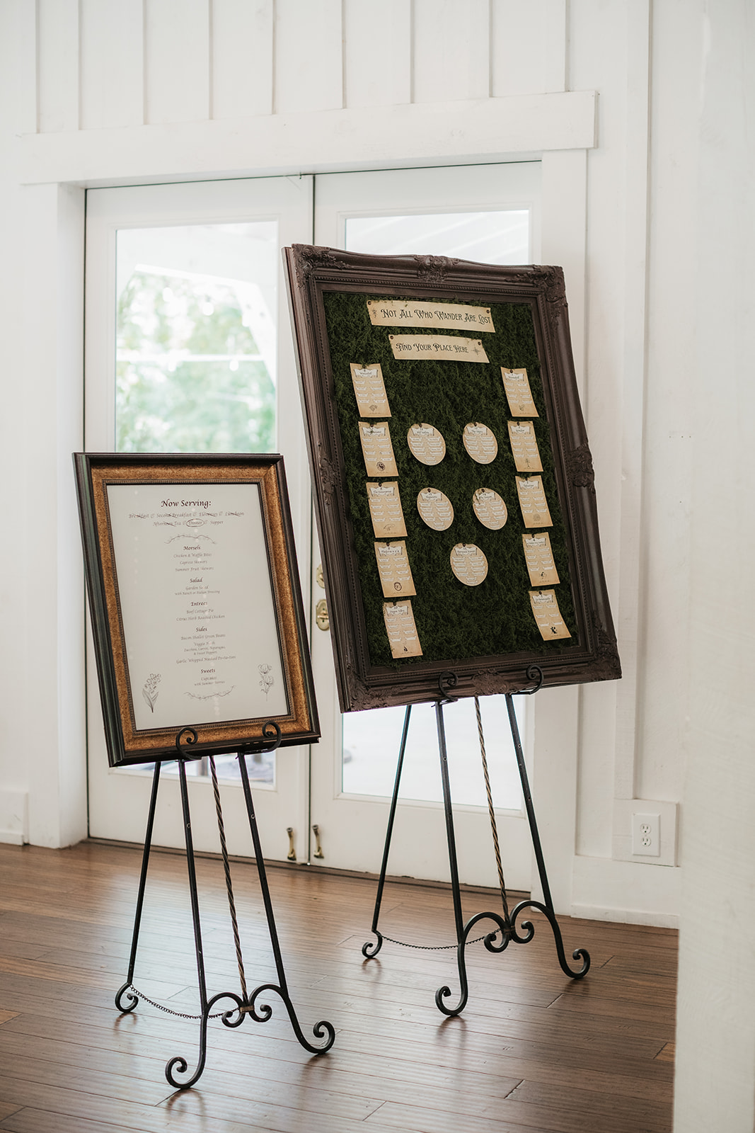 Two framed seating charts on metal easels, one with a list of names and the other displaying table arrangements on a green background, stand in front of glass doors for a wedding at Spain Ranch