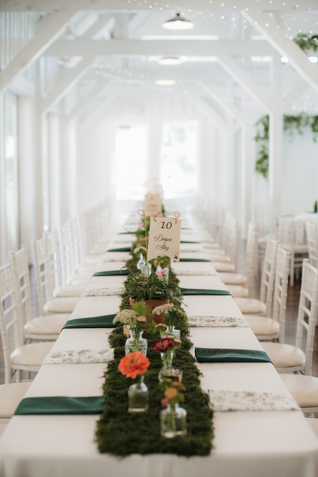 A long banquet table with white chairs is decorated with a green moss runner, small vases of flowers, and table number cards in a bright, white room for a wedding at spain ranch