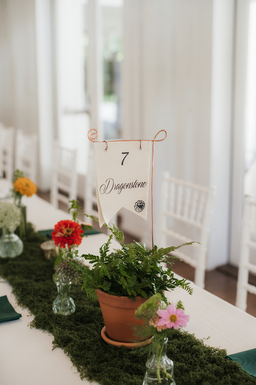 A long banquet table with white chairs is decorated with a green moss runner, small vases of flowers, and table number cards in a bright, white room for a wedding at spain ranch