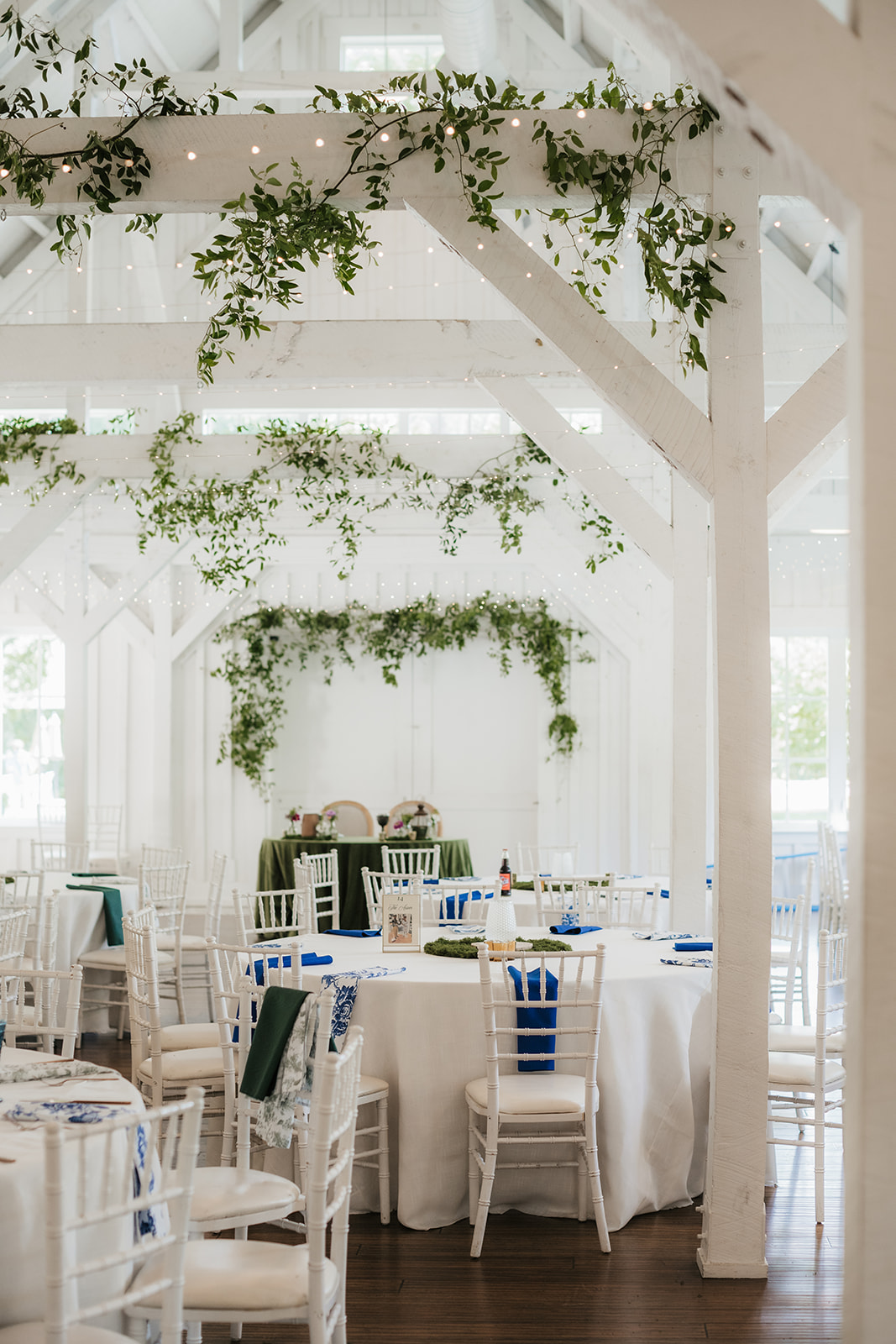 A long banquet table with white chairs is decorated with a green moss runner, small vases of flowers, and table number cards in a bright, white room for a wedding at spain ranch