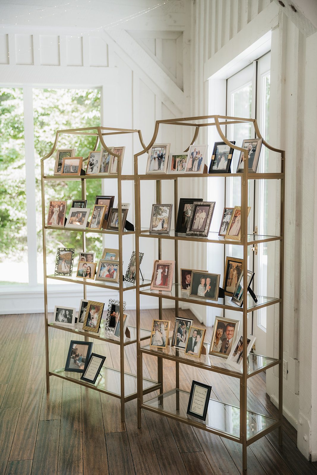 Two gold metal and glass shelves display numerous framed photographs in a well-lit room with large windows and wooden floors.