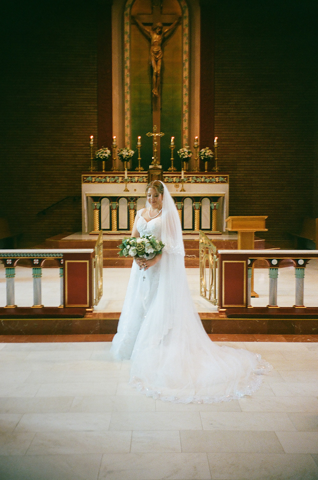 A bride in a white wedding dress and veil stands holding a bouquet in front of an altar inside a church.