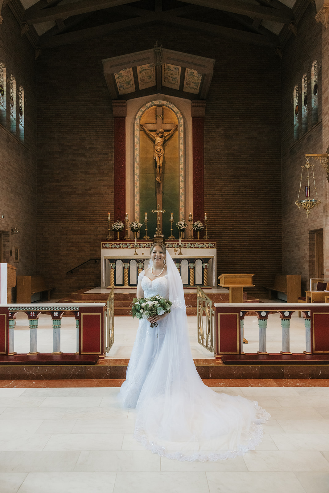 Catholic Wedding Photos of a bride and groom stand together in front of an altar inside a church, with the bride holding a bouquet and a large crucifix visible in the background.