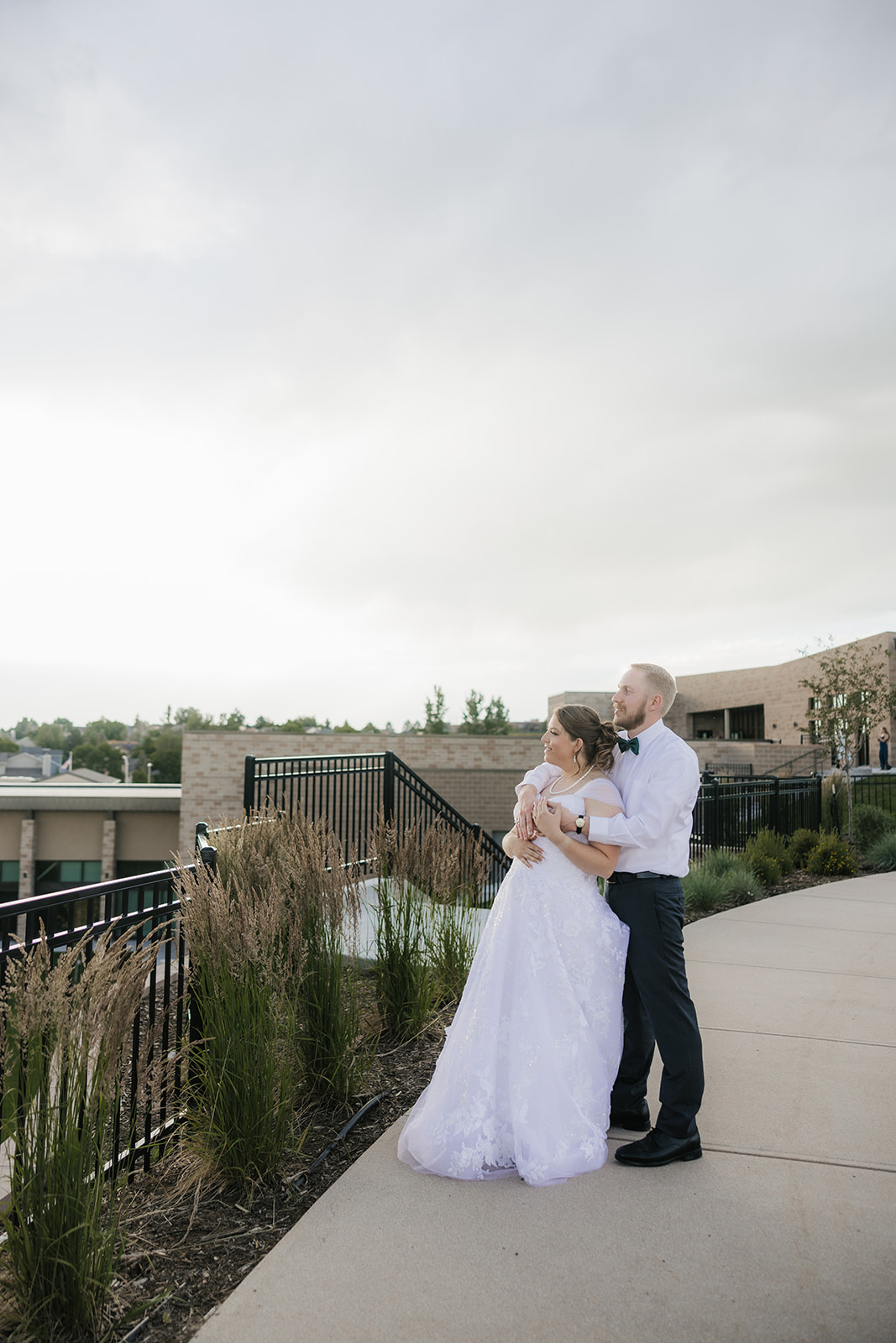 A bride and groom stand together outdoors on a walkway, looking into the distance beside a railing and tall plants under a cloudy sky.