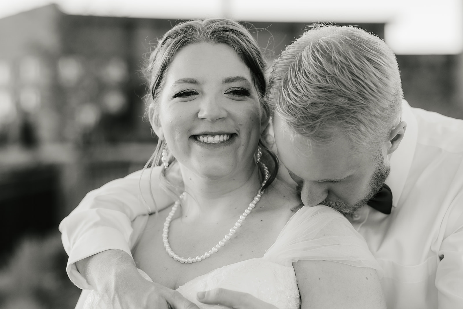 catholic wedding photos of A bride and groom share an embrace outdoors, with the groom wearing a shirt and bow tie and the bride in a wedding dress. The background features a brick wall and trees.