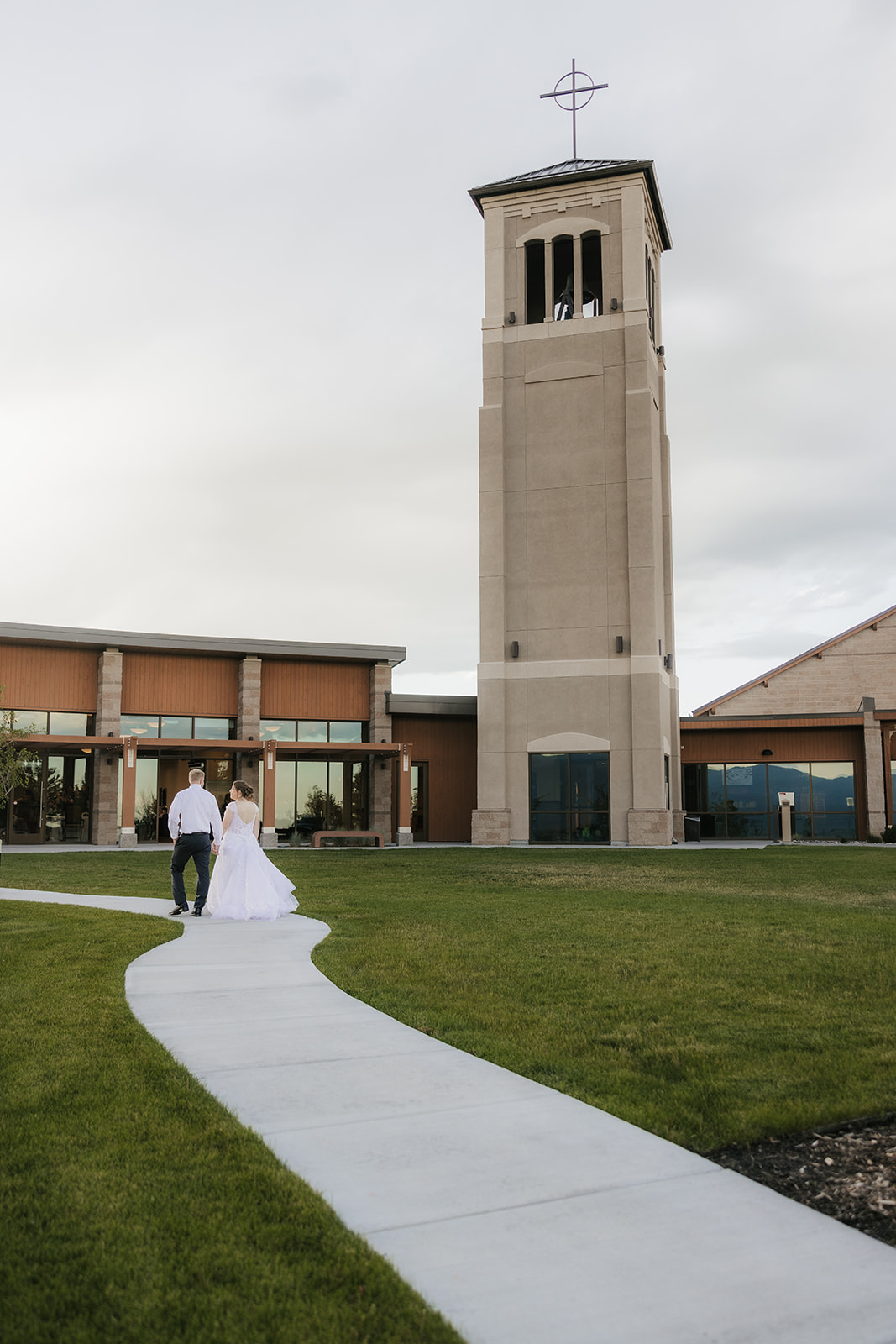 A bride and groom stand together outdoors on a walkway, looking into the distance beside a railing and tall plants under a cloudy sky.