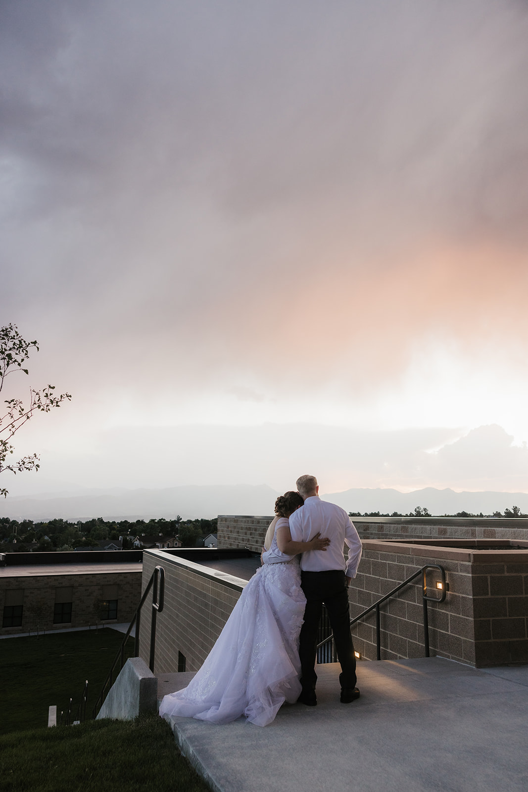 catholic wedding photos of A bride and groom kiss on an outdoor terrace with a scenic view of mountains, cloudy sky, and a town in the background.