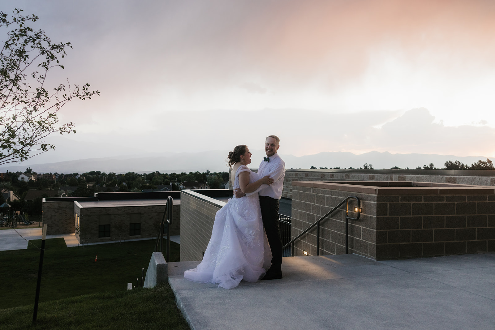 A bride and groom stand together outdoors on a walkway, looking into the distance beside a railing and tall plants under a cloudy sky.