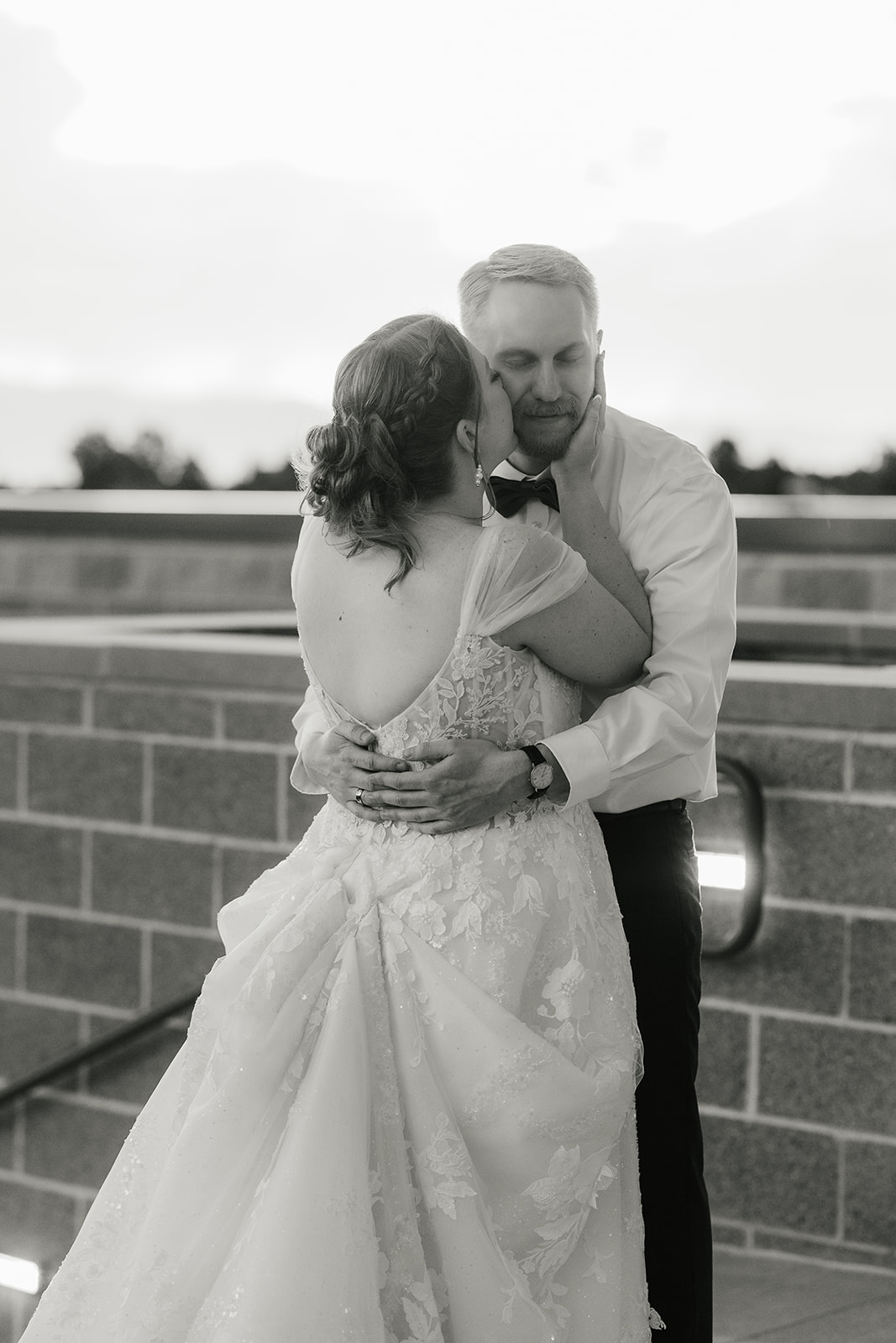 catholic wedding photos of A bride and groom share an embrace outdoors, with the groom wearing a shirt and bow tie and the bride in a wedding dress. The background features a brick wall and trees.
