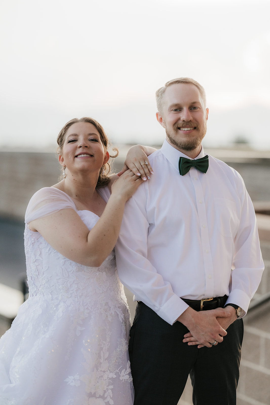 catholic wedding photos of A bride and groom share an embrace outdoors, with the groom wearing a shirt and bow tie and the bride in a wedding dress. The background features a brick wall and trees.