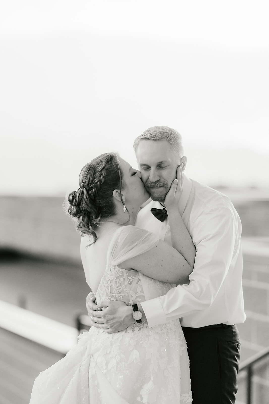 catholic wedding photos of A bride and groom share an embrace outdoors, with the groom wearing a shirt and bow tie and the bride in a wedding dress. The background features a brick wall and trees.