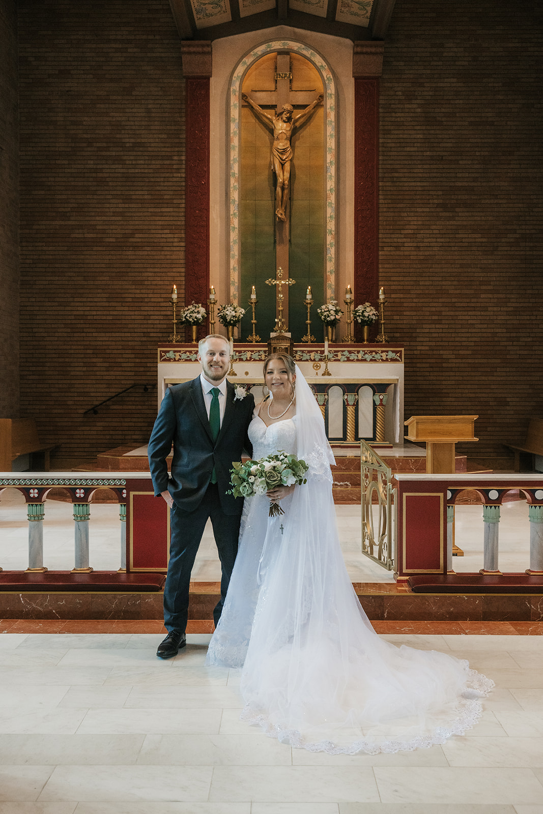 Catholic Wedding Photos of a bride and groom stand together in front of an altar inside a church, with the bride holding a bouquet and a large crucifix visible in the background.