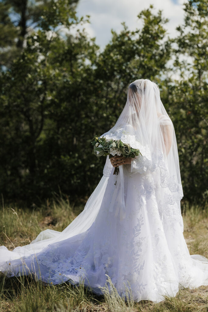 A bride and groom stand outdoors in wedding attire, facing each other under a veil, surrounded by greenery and a cloudy sky for catholic wedding photos