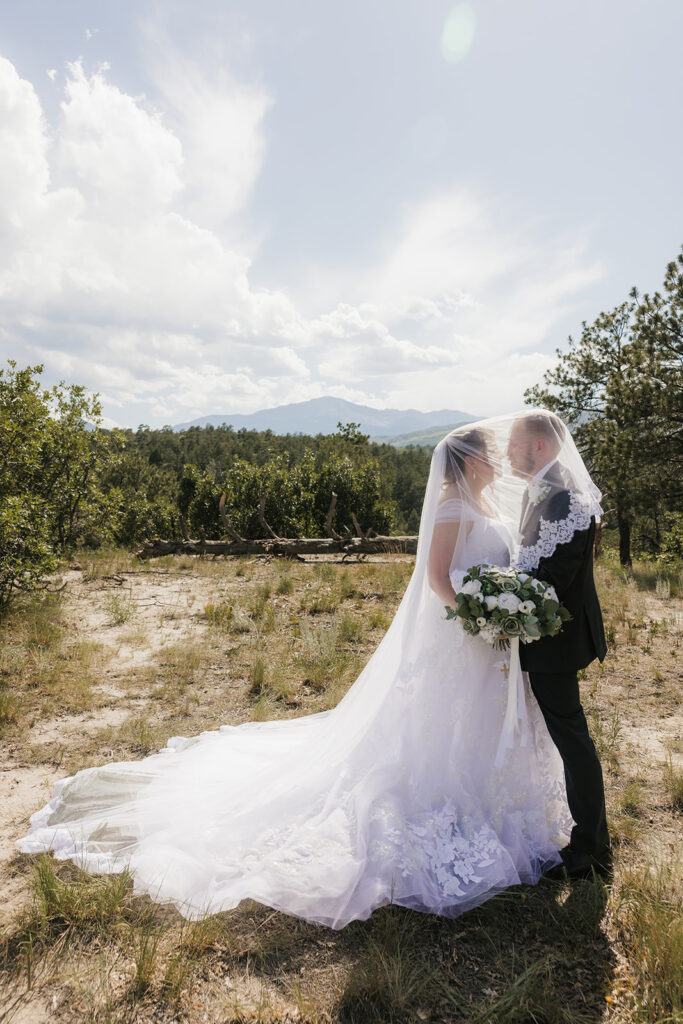 A bride and groom stand outdoors in wedding attire, facing each other under a veil, surrounded by greenery and a cloudy sky for catholic wedding photos