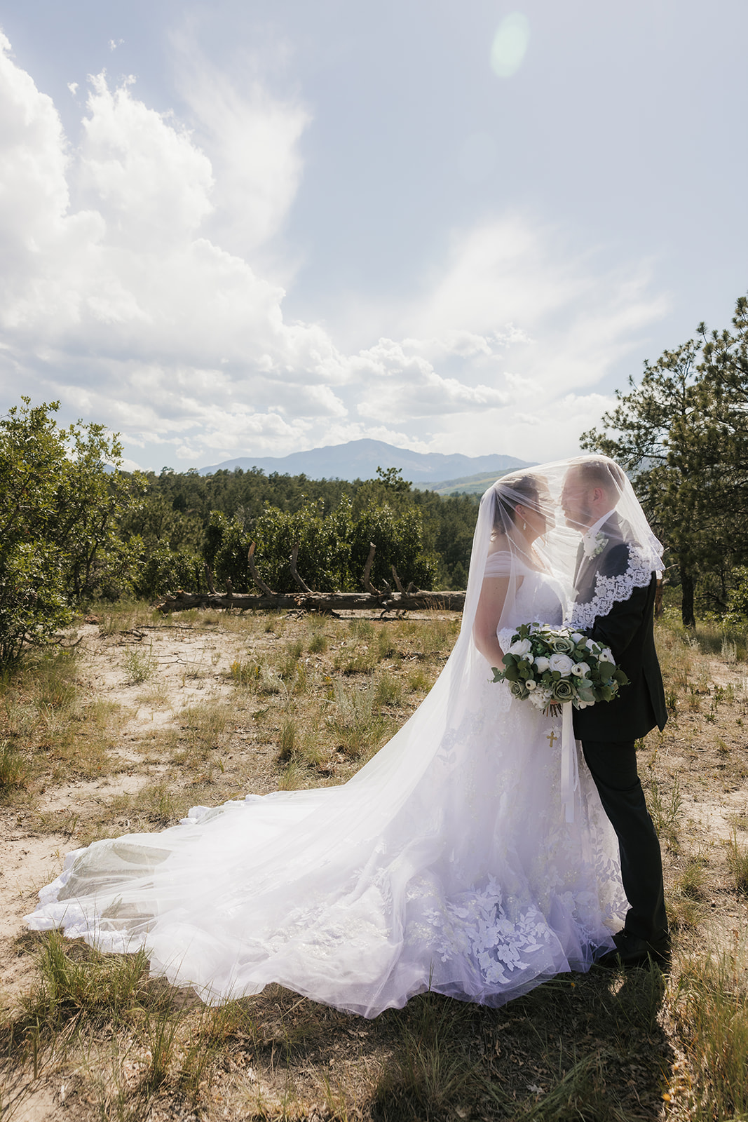 A bride and groom stand outdoors in wedding attire, facing each other under a veil, surrounded by greenery and a cloudy sky for catholic wedding photos