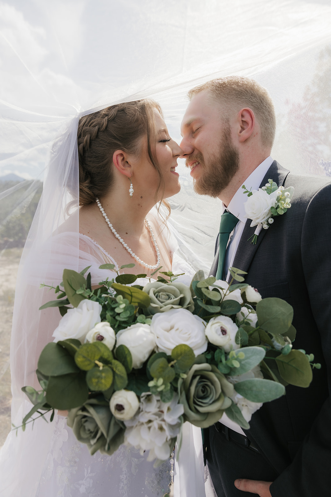 catholic wedding photos of A bride and groom stand outdoors in wedding attire, facing each other under a veil, surrounded by greenery and a cloudy sky for catholic wedding photos