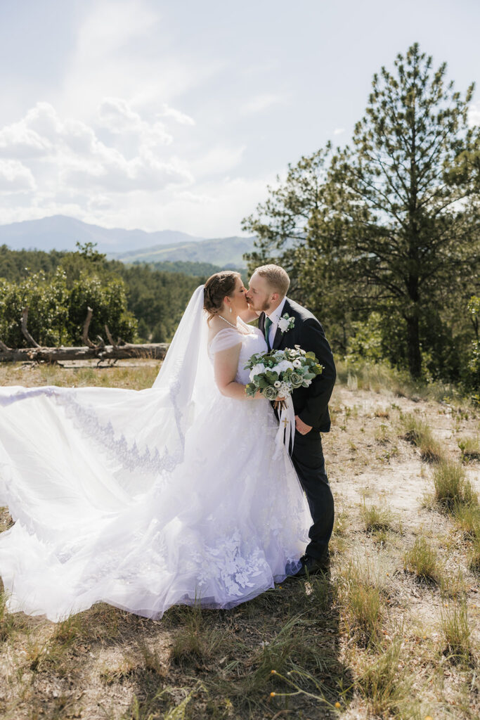 A bride and groom stand outdoors in wedding attire, facing each other under a veil, surrounded by greenery and a cloudy sky for catholic wedding photos