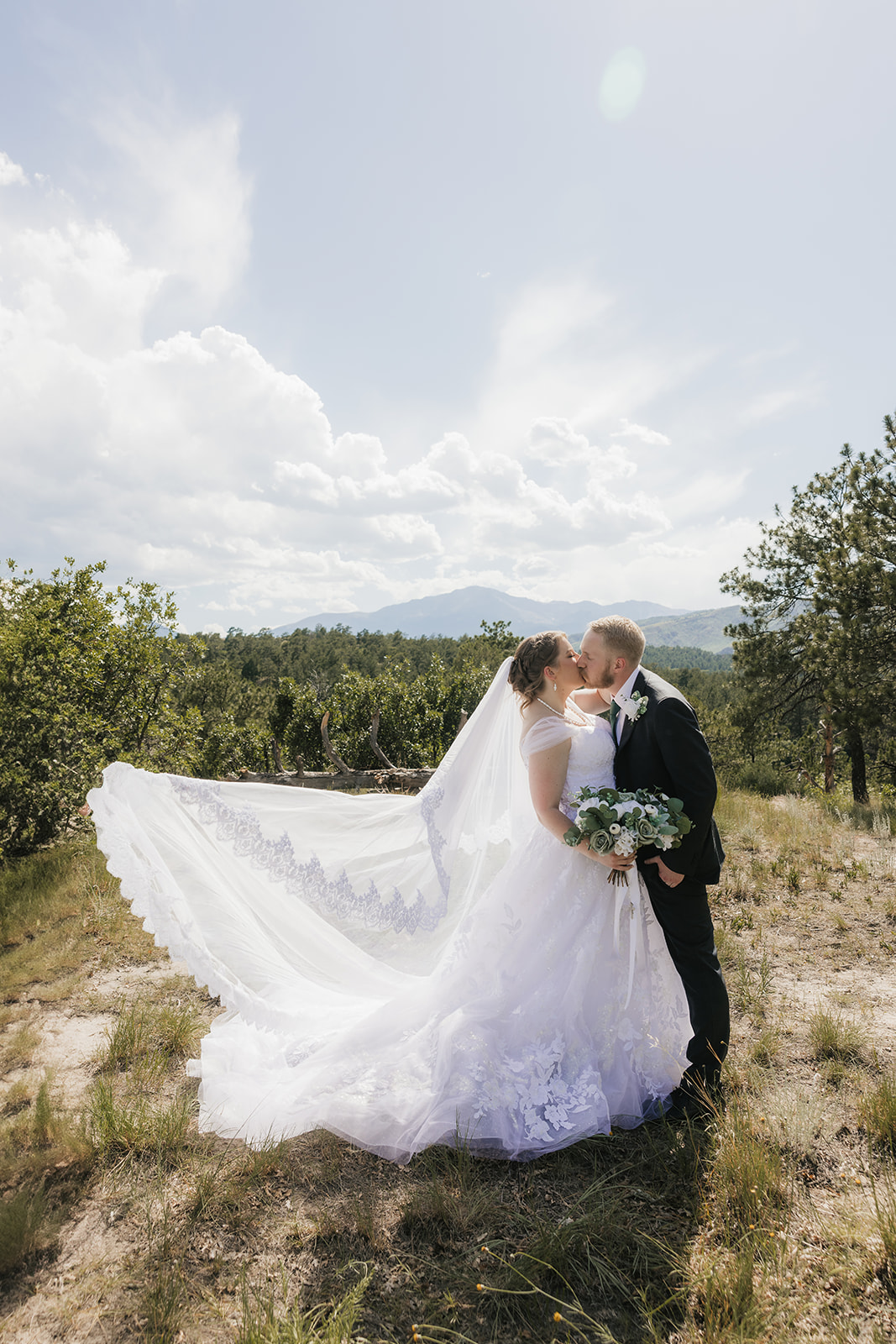 catholic wedding photos of A bride and groom stand outdoors in wedding attire, facing each other under a veil, surrounded by greenery and a cloudy sky for catholic wedding photos