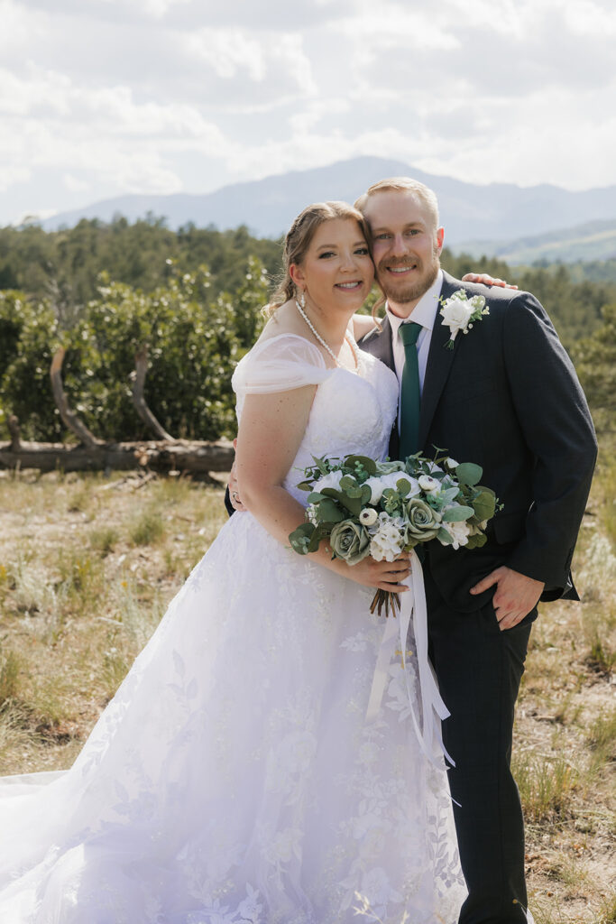 A bride and groom stand outdoors in wedding attire, facing each other under a veil, surrounded by greenery and a cloudy sky for catholic wedding photos