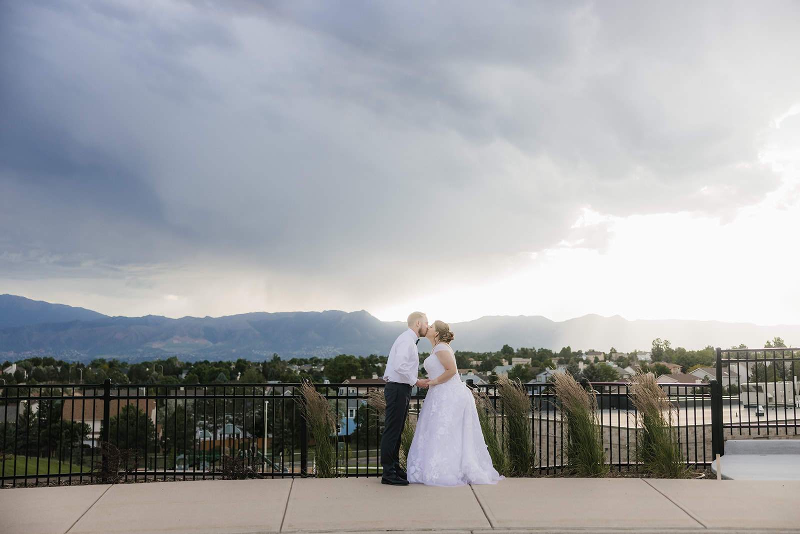 catholic wedding photos of A bride and groom kiss on an outdoor terrace with a scenic view of mountains, cloudy sky, and a town in the background.