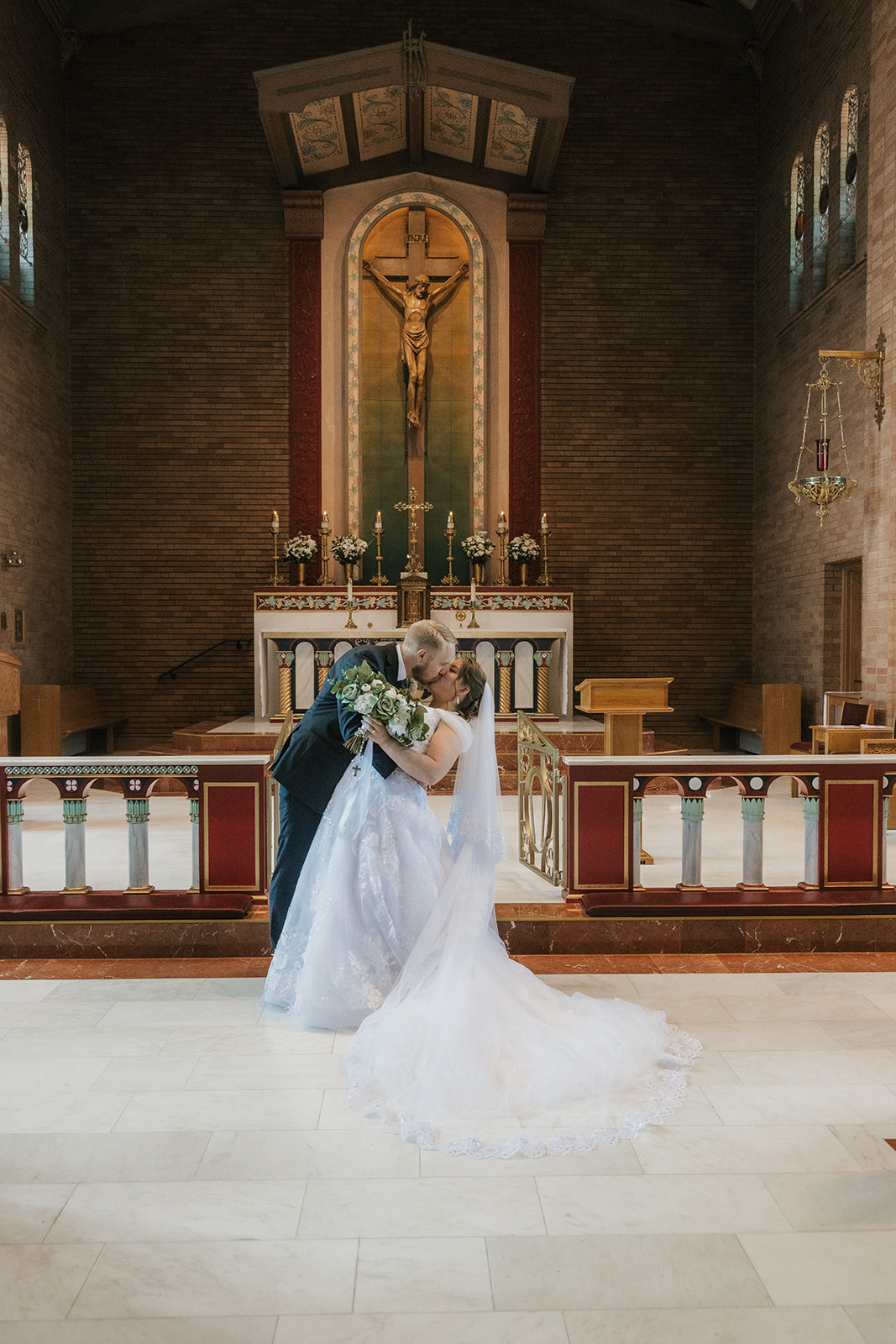 Catholic Wedding Photos of a bride and groom stand together in front of an altar inside a church, with the bride holding a bouquet and a large crucifix visible in the background.