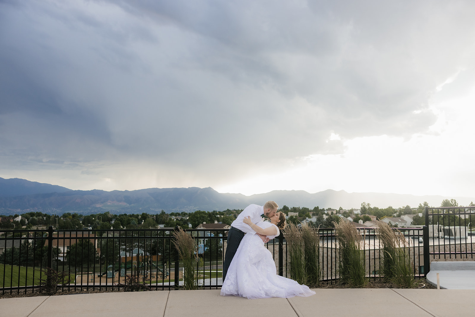 catholic wedding photos of A bride and groom kiss on an outdoor terrace with a scenic view of mountains, cloudy sky, and a town in the background.
