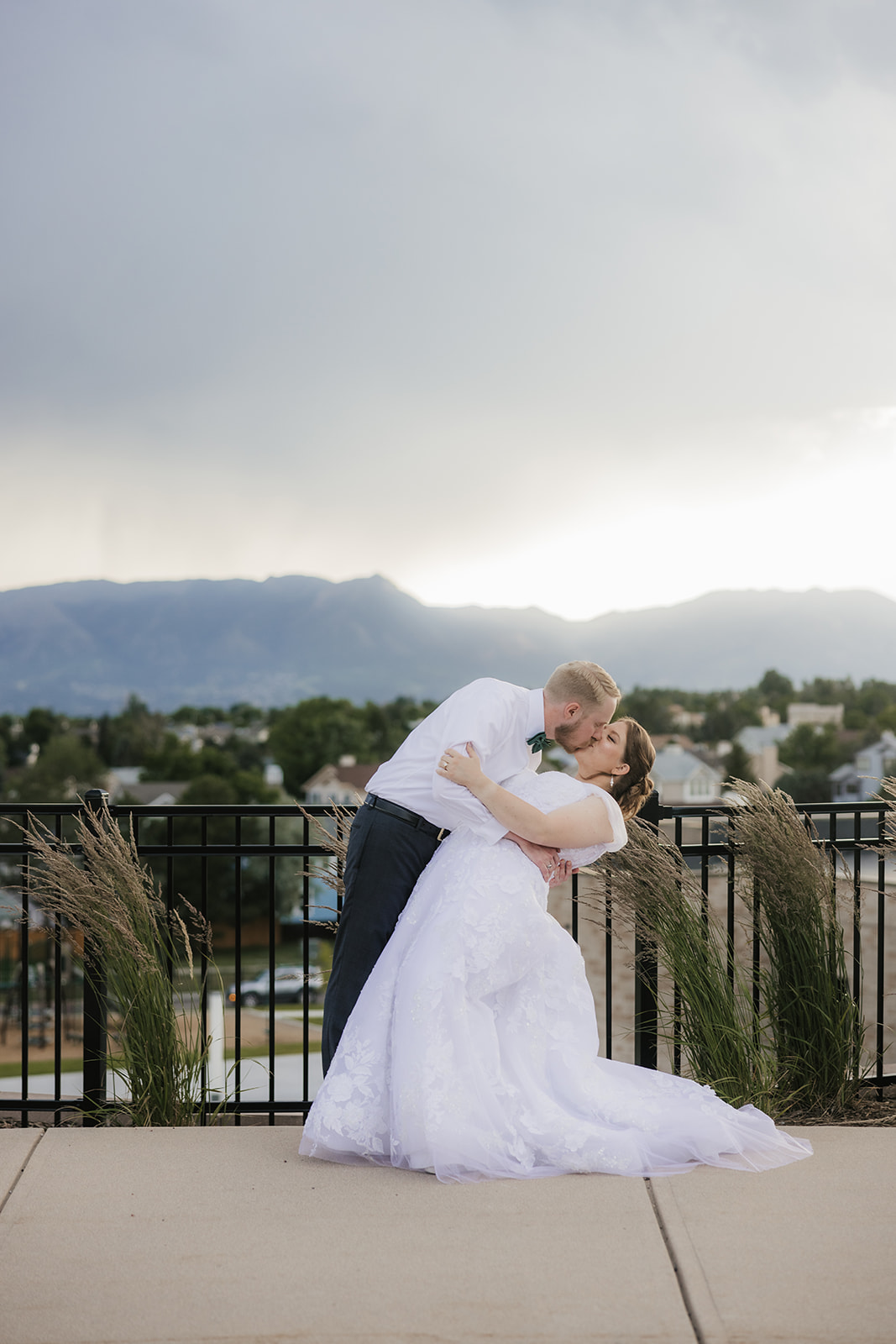 catholic wedding photos of A bride and groom kiss on an outdoor terrace with a scenic view of mountains, cloudy sky, and a town in the background.