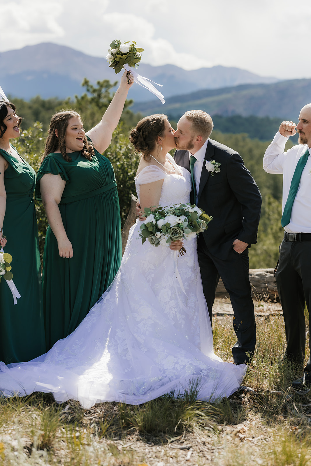 catholic wedding photos of a bride and groom stand outdoors with their wedding party, dressed in green and white, posing for a photo against a natural landscape with trees and mountains.
