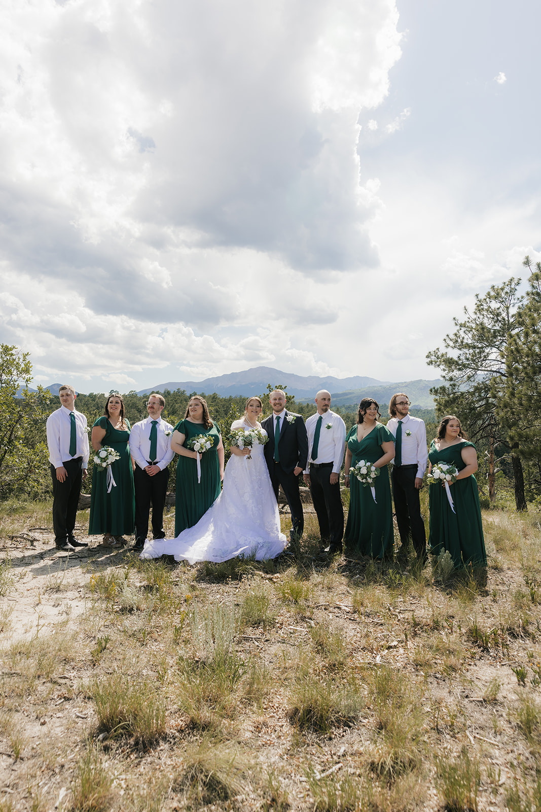 catholic wedding photos of a bride and groom stand outdoors with their wedding party, dressed in green and white, posing for a photo against a natural landscape with trees and mountains.
