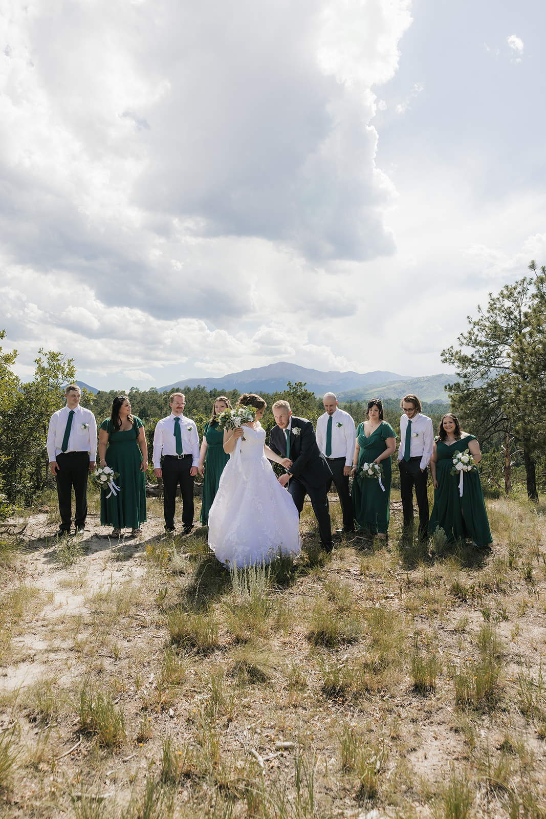 catholic wedding photos of a bride and groom stand outdoors with their wedding party, dressed in green and white, posing for a photo against a natural landscape with trees and mountains.