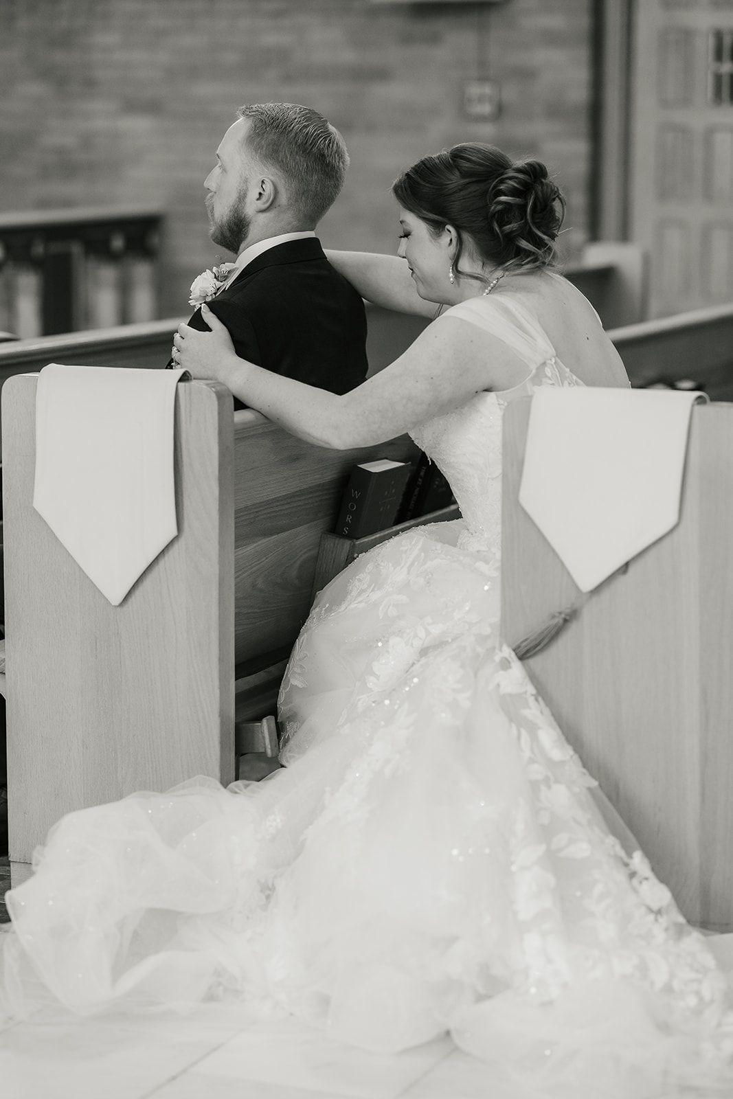 A bride and groom sit together in a church pew, facing the altar; the bride’s dress train spreads across the floor.